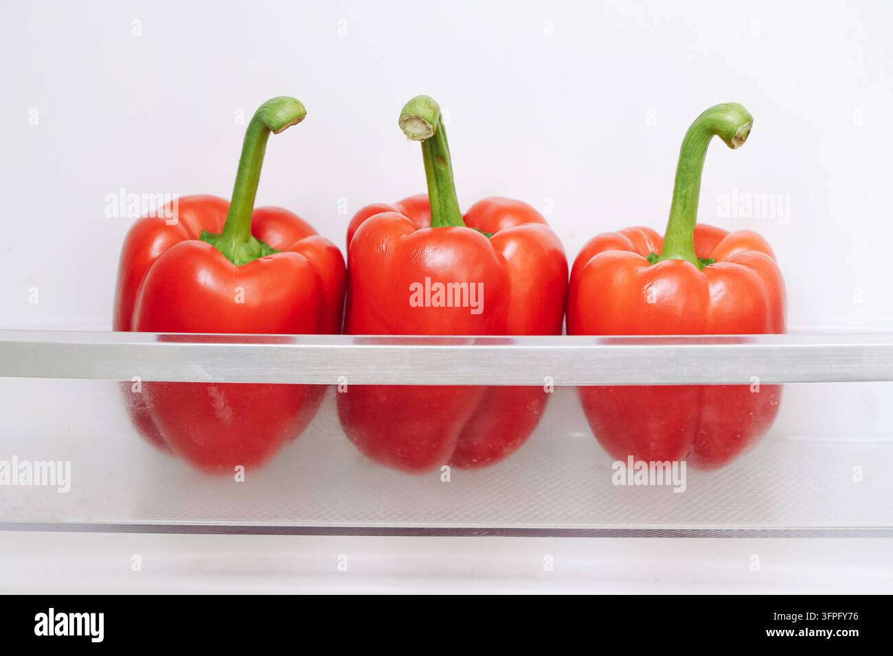 Drei frische rote Paprikaschoten in der durchsichtigen Kühlschank-Schublade. Hintergrund für die Lebensmittelaufbewahrung von Gemüse, Frische, gesundes Essen und die heimische Küche. Stockfoto