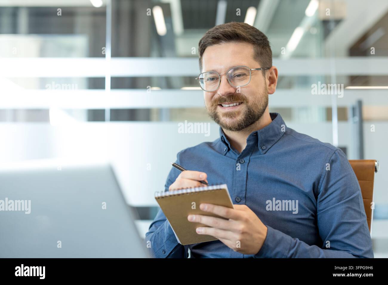 Junger lächelnder Geschäftsmann mit Brille, der im Büro arbeitet, ein Notizbuch und einen Stift hält und auf einen Laptop-Bildschirm schaut, während er an einem Online-Meeting oder Webinar teilnimmt Stockfoto