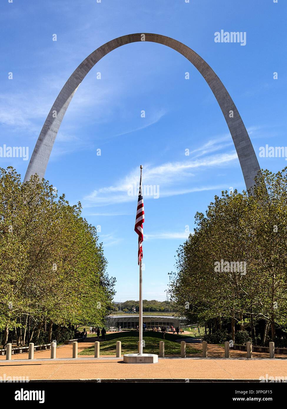 Das ikonische Gateway Arch erhebt sich vor einem klaren blauen Himmel in St. Louis, Missouri, eingerahmt von Bäumen mit einer wehenden amerikanischen Flagge im Vordergrund. Besucher Stockfoto