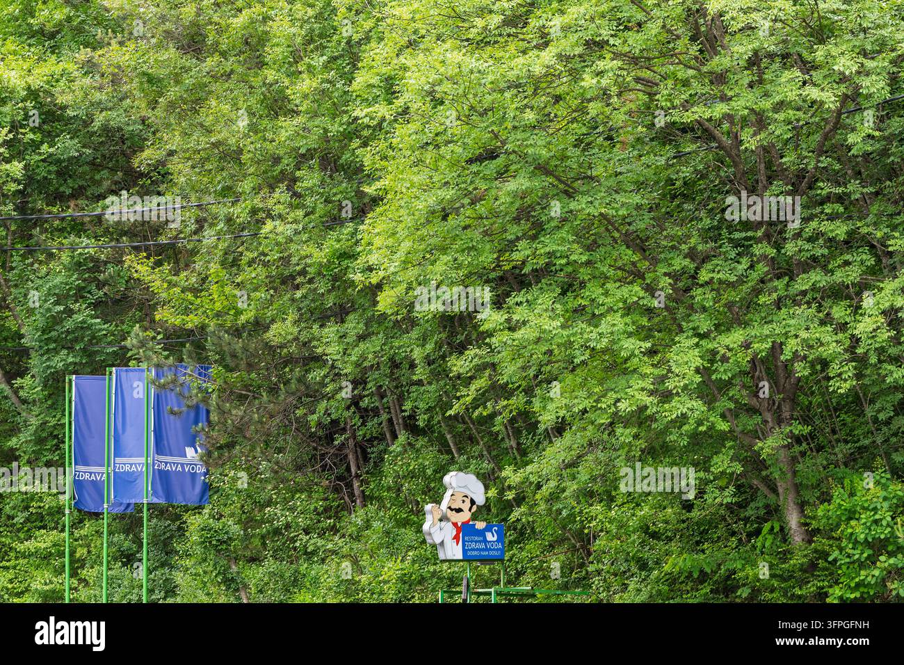 Jablanica, Bosnien und Herzegowina - 17. Mai 2026: Blaue Werbefahnen und ein Cartoon-Koch-Schild werben für einen gesunden Wasserstand vor einem dichten, belaubten F Stockfoto