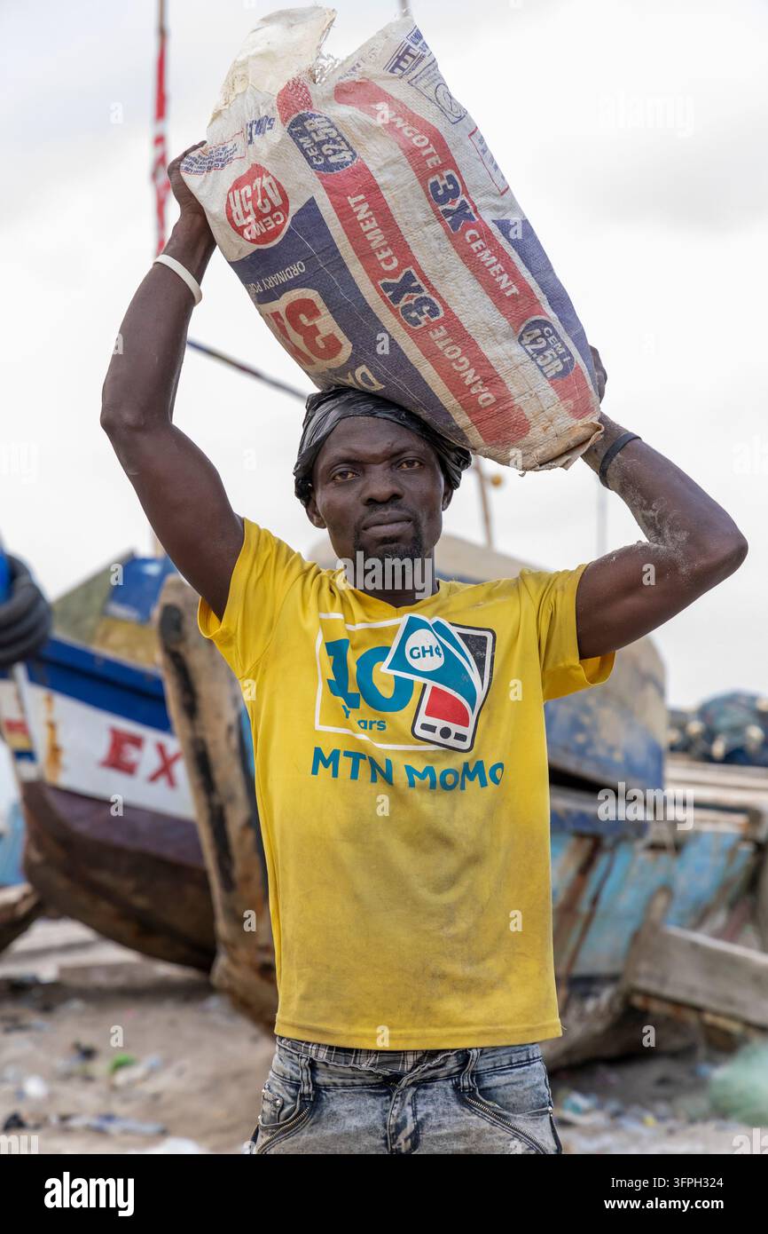 Ein Mann trägt einen schweren Zementbeutel auf seinem Kopf in der Nähe von gestrandeten Fischerbooten, Zentralregion, Cape Coast, Ghana Stockfoto