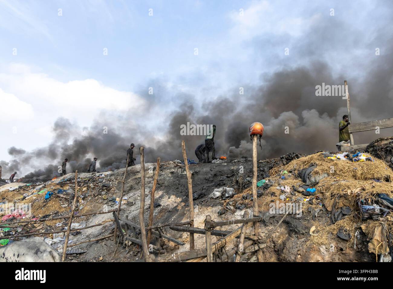 Arbeiter stehen auf einem Hügel aus brennendem Müll unter dickem schwarzem Rauch mit einem Motorradhelm, der auf einem Holzstab lehnt, Greater Accra Region, Accra, Gh Stockfoto
