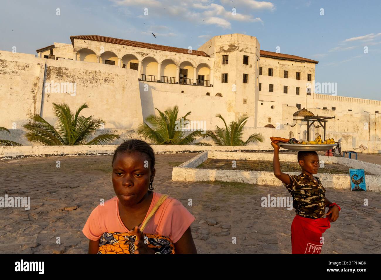Frauen, die außerhalb der Festung Elmina spazieren, eine trägt ein Tablett mit Essen auf dem Kopf, Zentralregion, Elmina, Ghana Stockfoto