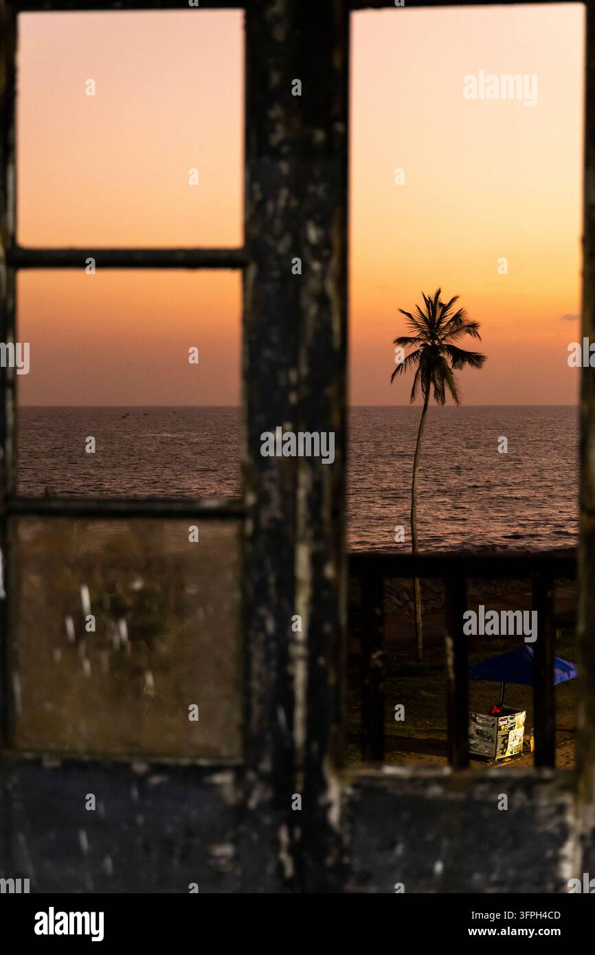 Silhouette einer Palme, die durch einen alten hölzernen Fensterrahmen bei Sonnenuntergang gesehen wird, Zentralregion, Elmina, Ghana Stockfoto