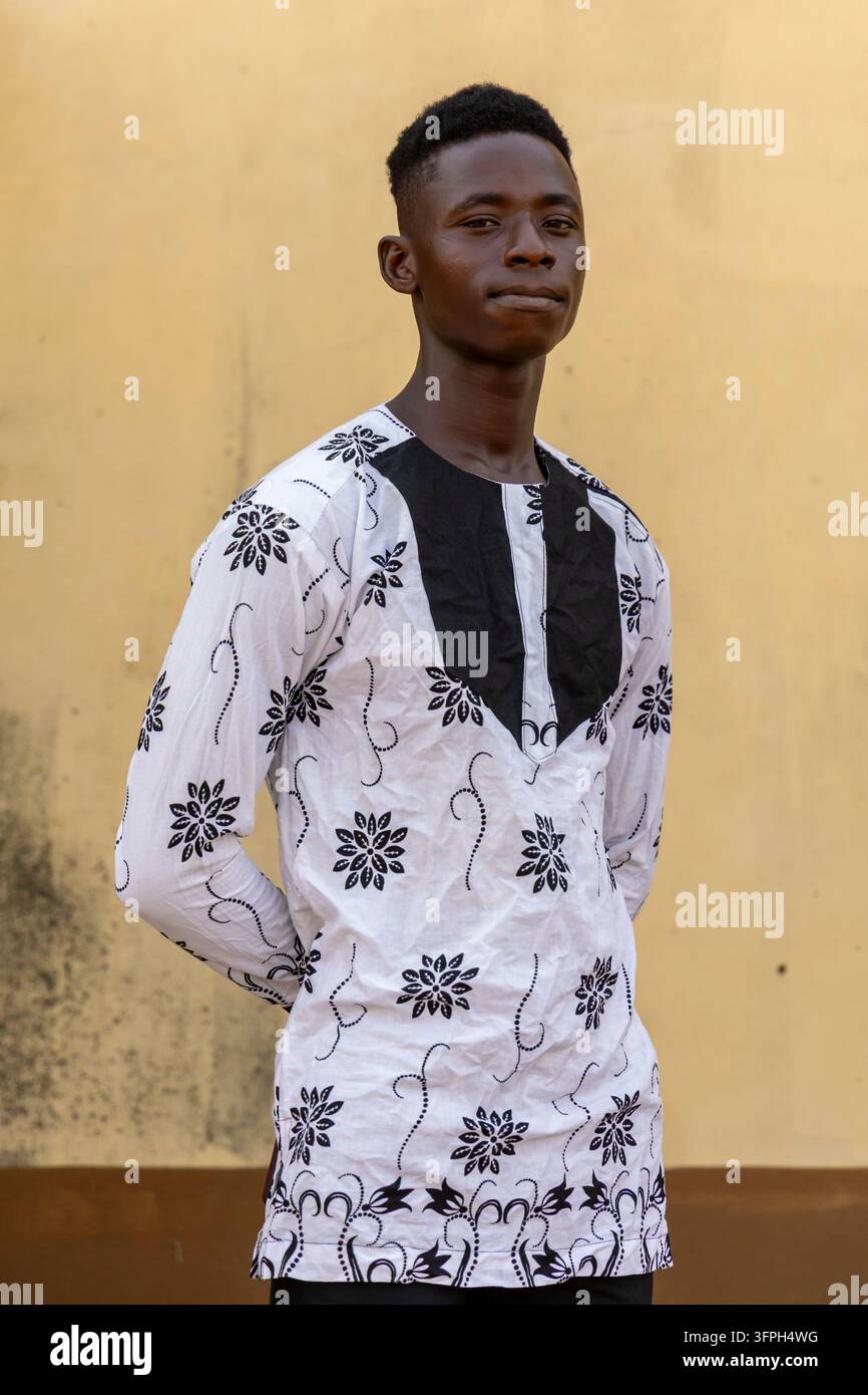 Porträt eines jungen Mannes, der ein weißes traditionelles Hemd mit schwarzem Blumenmuster vor einer gelben Wand trägt, Zentralregion, Elmina, Ghana Stockfoto
