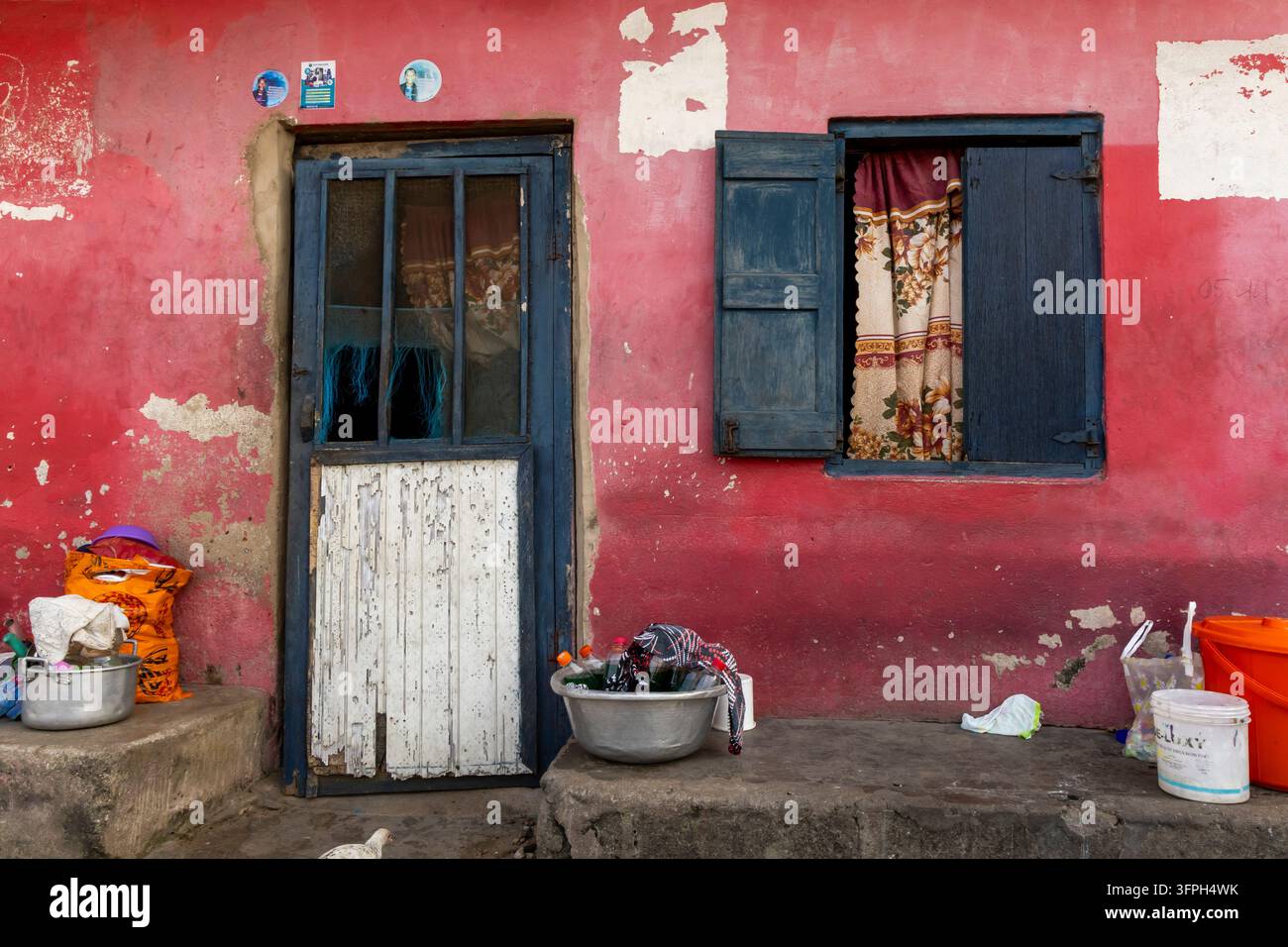 Verwitterte rote Hausfassade mit einer blauen Tür und einem verhangenen Fenster, umgeben von Becken und Eimern, Zentralregion, Elmina, Ghana Stockfoto