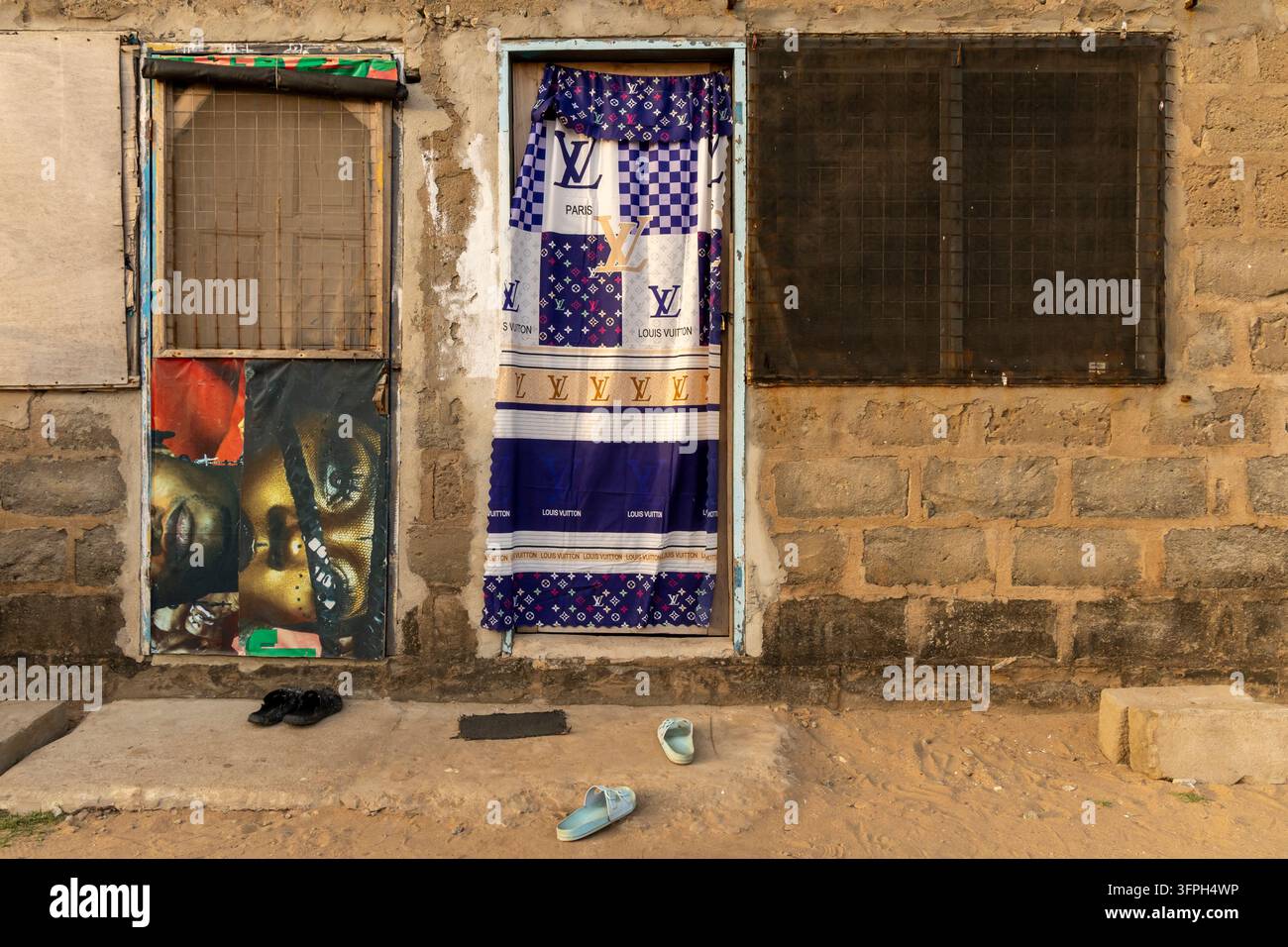 Eingang eines Zementblockhauses mit einem Vorhang aus Monogrammdruckstoff und Sandalen, die auf dem sandigen Boden liegen, Zentralregion, Elmina, Ghana Stockfoto