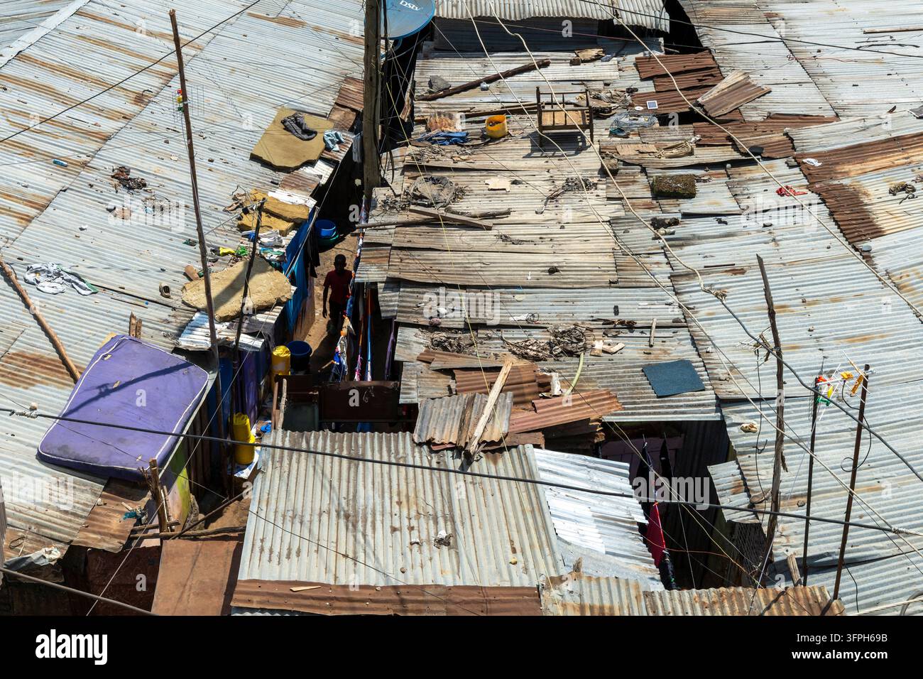 Luftaufnahme von rostigen Wellblechdächern, die dicht gedrängt im Kibera-Gebiet stehen, mit einem Jungen, der in einer schmalen Gasse darunter geht, und Satellitenschüssel und Drähten Stockfoto
