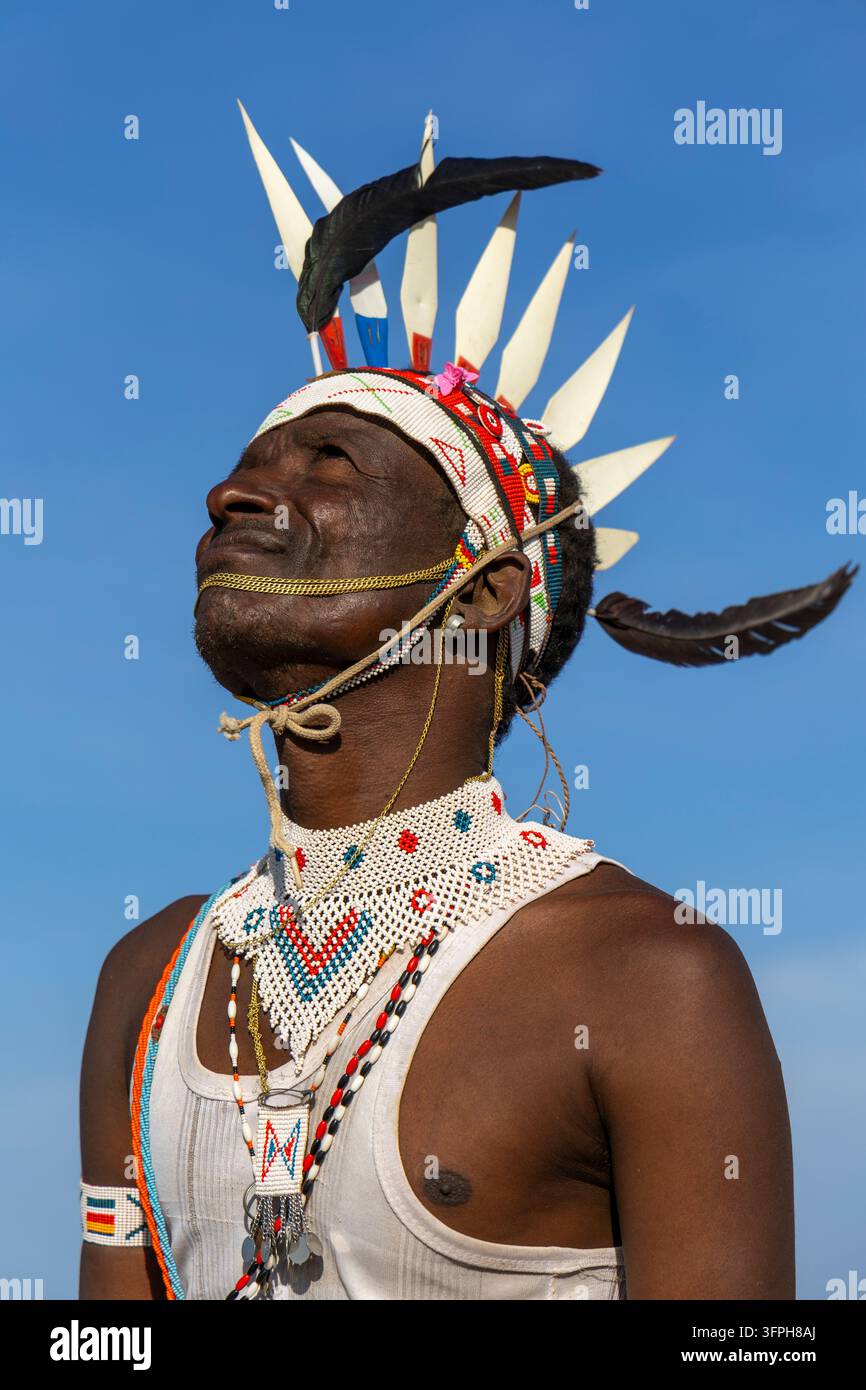 Mann des Rendille-Stammes schaut nach oben und trägt ein mit Perlen verziertes Stirnband mit weißen Stacheln und einer schwarzen Feder, einen mit Perlen verzierten Kragen und ein weißes Tanktop vor blauem Himmel, M Stockfoto