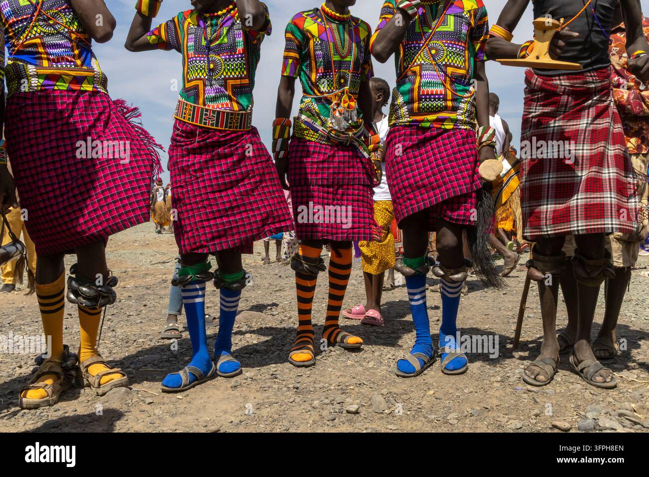 Niedrigwinkelaufnahme von Männern des Turkana-Stammes in bunten bedruckten Hemden und karierten Kilts mit Knöchelrasseln und gestreiften Socken am Lake Turkana Cultural Fes Stockfoto