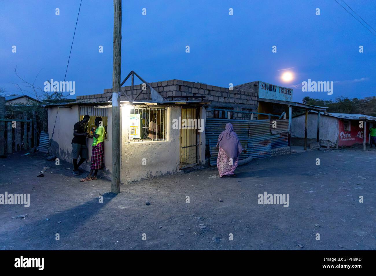 Straßenladen namens Shaaban Traders bei Einbruch der Dunkelheit unter vollem Mond beleuchtet, mit zwei Männern, die am Fenster lehnen, und einer Frau, die vorbeigeht, Marsabit County, Loiyan Stockfoto