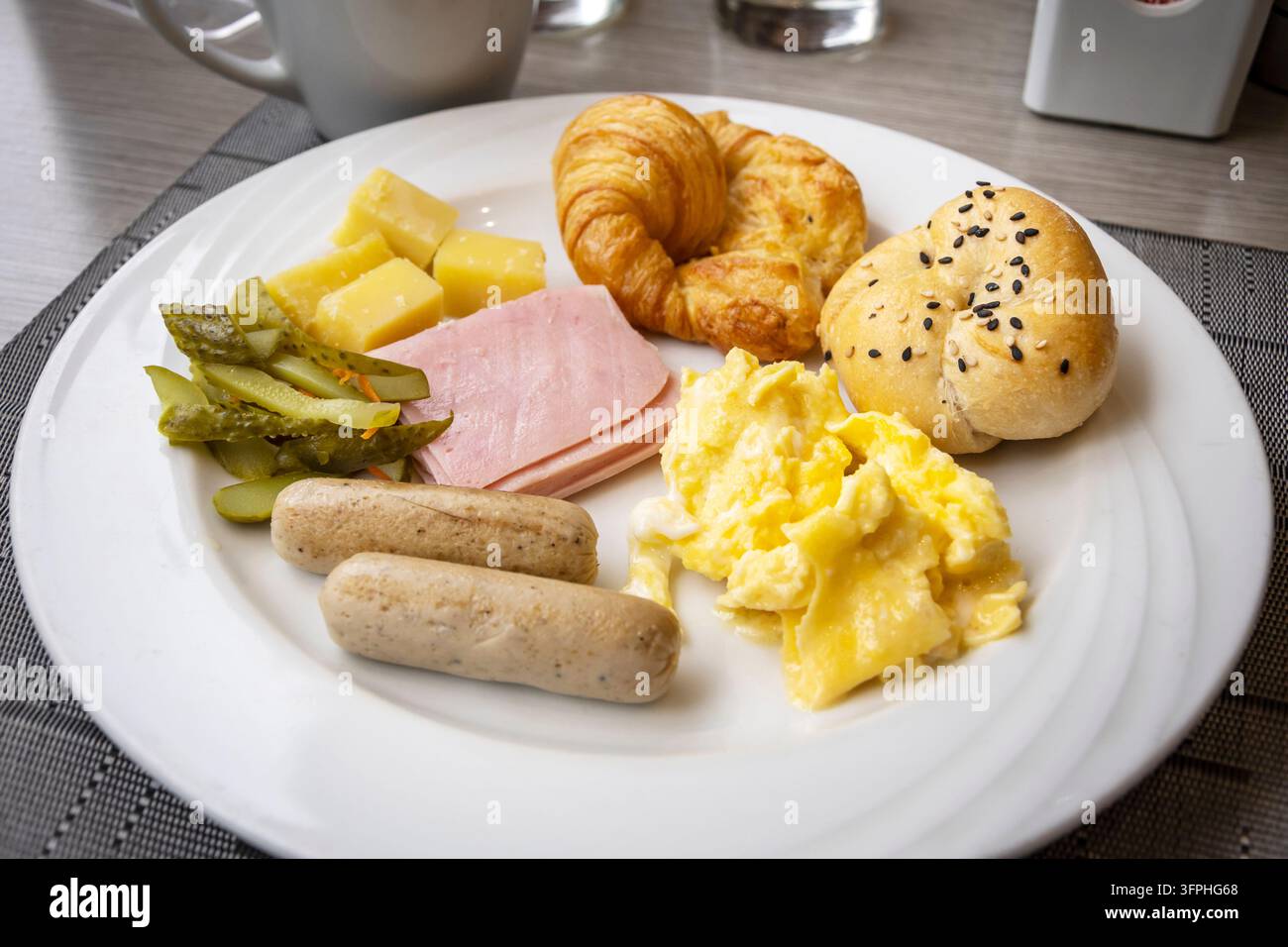 Ein herzhaftes Frühstücksteller mit Rührei, Schinken, Würstchen, Croissant und frischem Brot Stockfoto