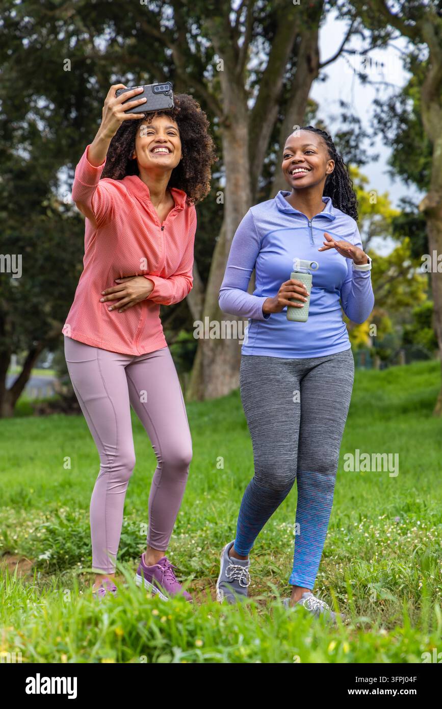 Verschiedene Freundinnen gehen im Park in Sportkleidung und machen mit dem Handy ein Selfie mit wiederverwendbarer Flasche. Draußen, Grünflächen, Bäume, Fitness, Hydration, Natur, lässig Stockfoto