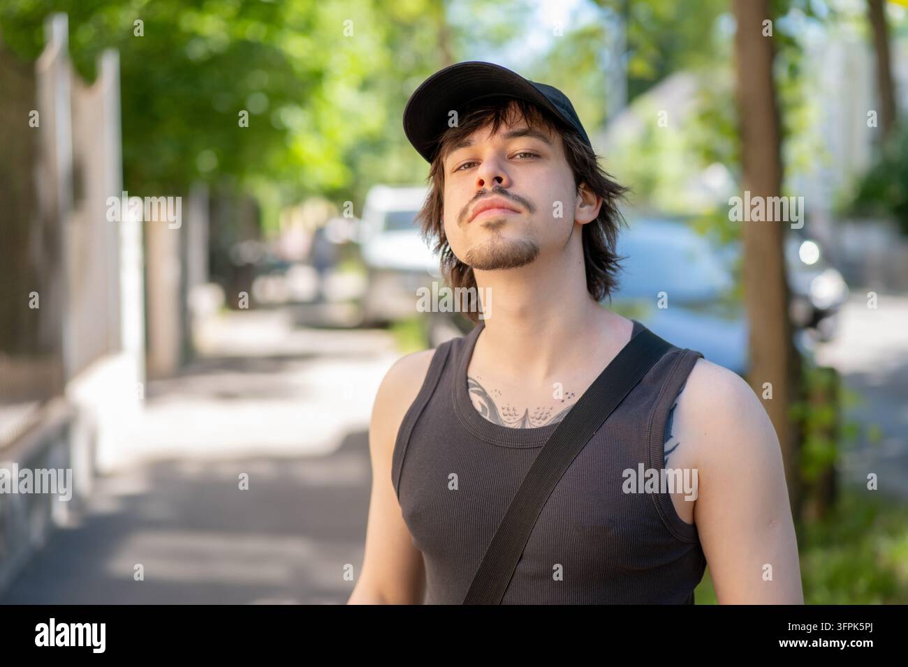 Ein junger Mann mit einer schwarzen Kappe und einem Tanktop posiert an einem sonnigen Stadtrandstein. Stockfoto