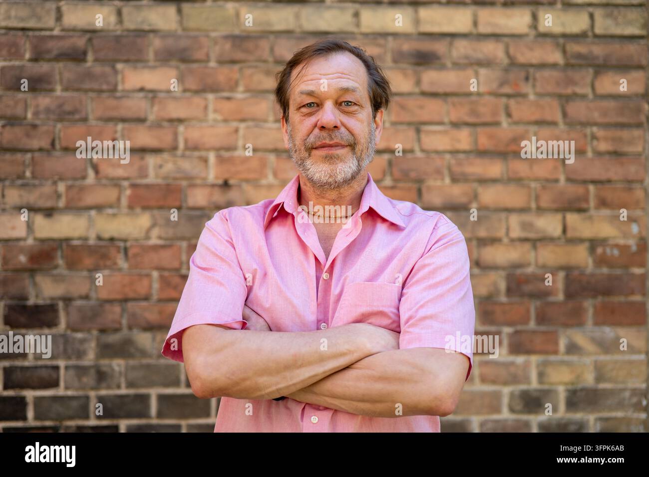 Ein mittelalter Mann in einem rosa Hemd steht mit verschränkten Armen vor einer Ziegelmauer. Stockfoto