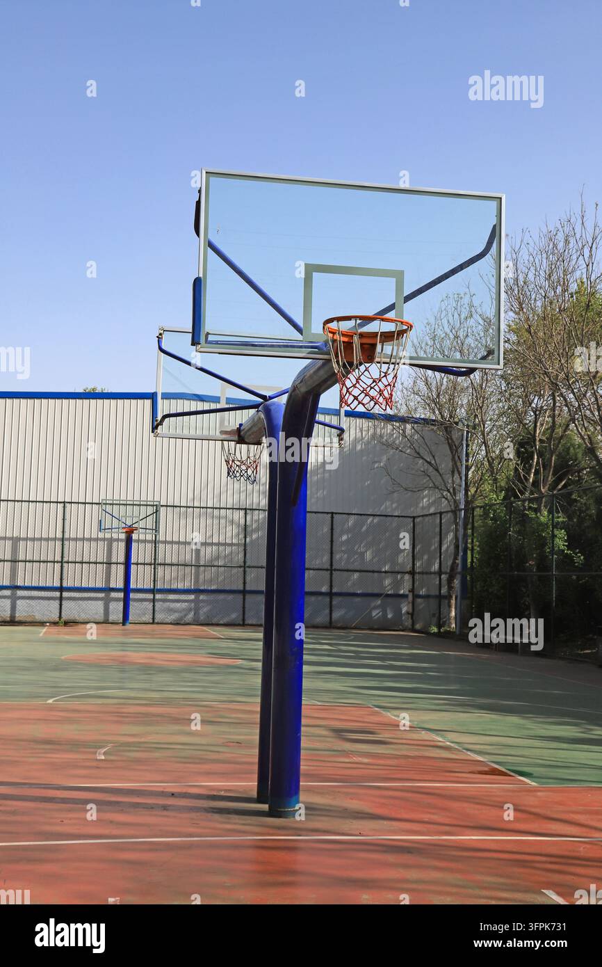 An einem sonnigen Tag auf einem Außenspielfeld steht ein Basketballkorb mit einem blauen Pfosten auffällig im Blickfeld. Das transparente Rückboard und der orangefarbene Ring sind klar Stockfoto