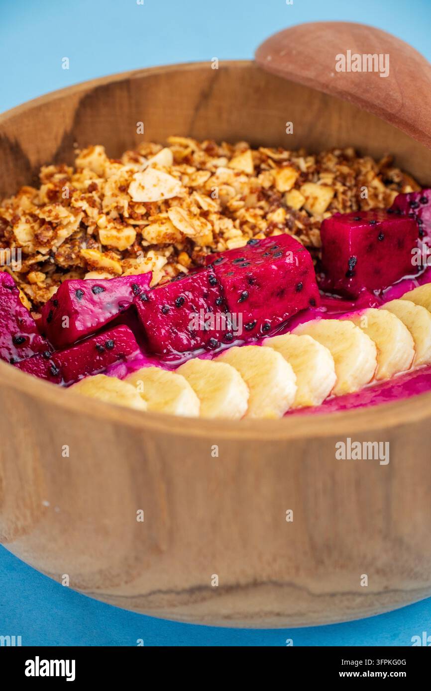 Pitaya-Smoothie-Schale mit geschnittener Banane und Granola, serviert in einer Holzschale auf blauem Hintergrund. Stockfoto