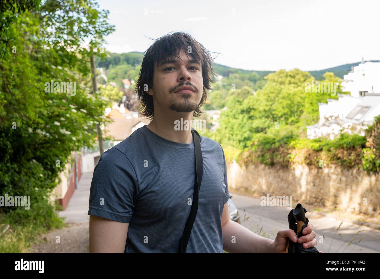 Ein junger Mann steht auf einer ruhigen Hügelstraße mit Bäumen und Häusern im Hintergrund. Stockfoto
