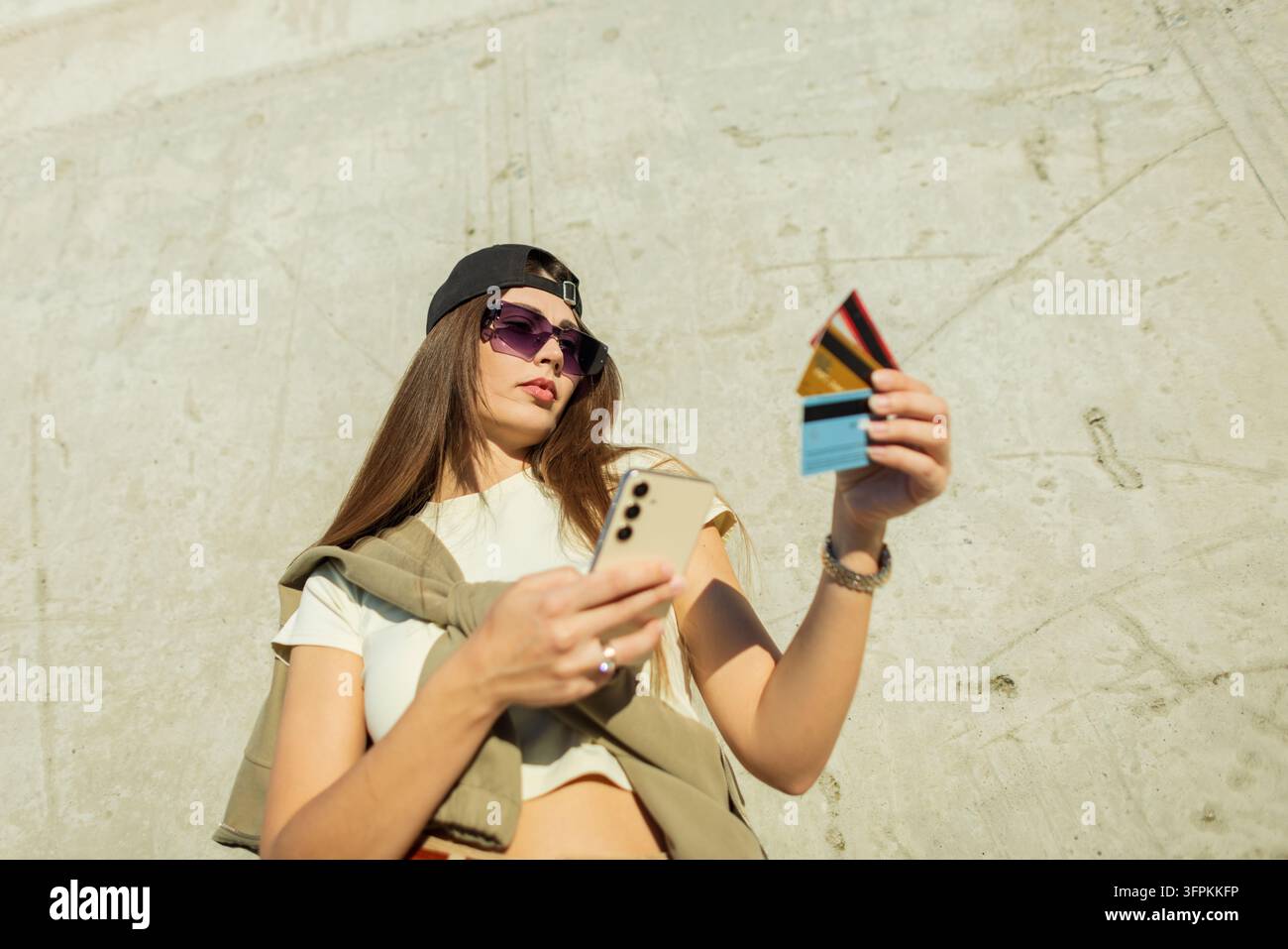 junge stilvolle Frau tätigt einen Online-Einkauf mit ihrem Smartphone und mehreren Kreditkarten, aufgenommen aus einem dynamischen niedrigen Winkel vor einem texturierten urbanen Hintergrund Stockfoto