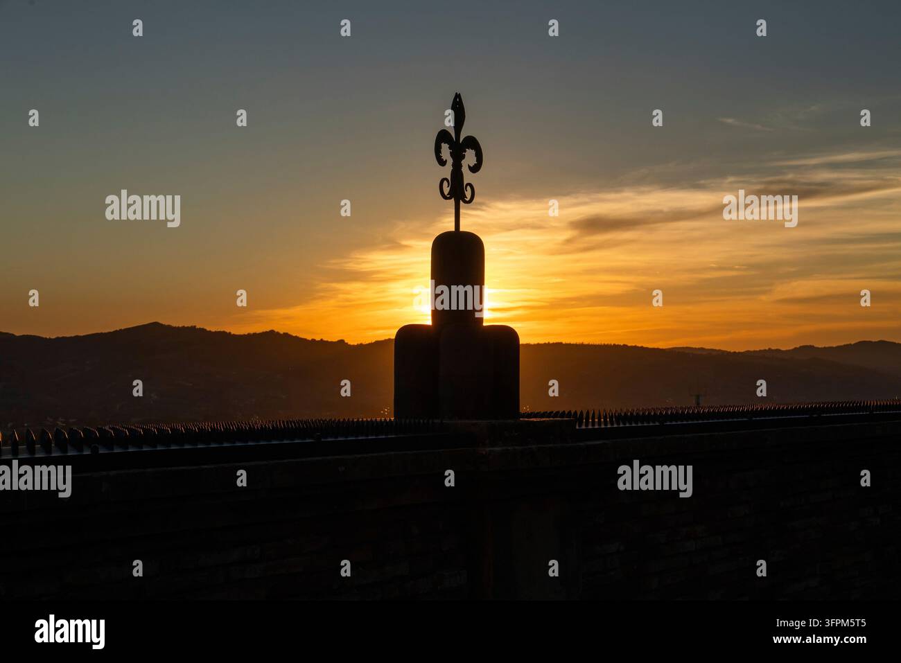 Ein Fleur-de-Lis-Symbol, stilisierte Lilie, hebt sich als dunkle Silhouette vor dem orangefarbenen Sonnenuntergang und einer Bergkette ab, Gubbio, Umbrien, Italien Stockfoto