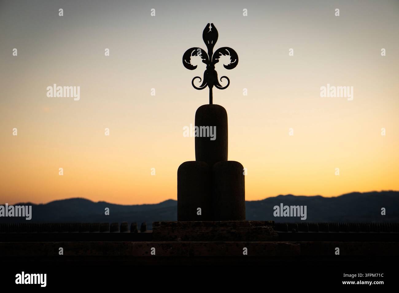 Ein Fleur-de-Lis-Symbol, stilisierte Lilie, hebt sich als dunkle Silhouette vor dem orangefarbenen Sonnenuntergang und einer Bergkette ab, Gubbio, Umbrien, Italien Stockfoto