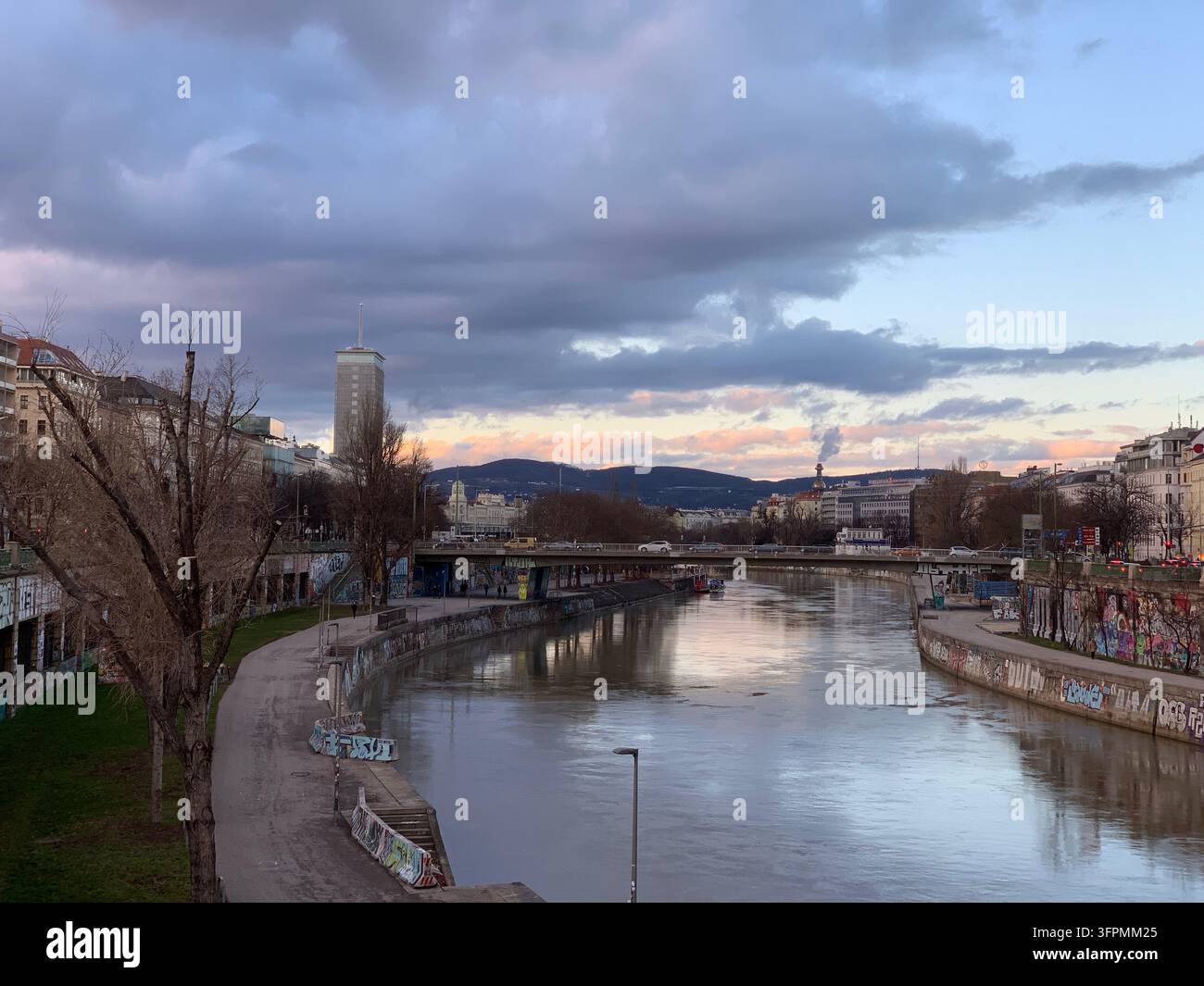 Donau in Wien, Österreich, mit der Stadtlandschaft auf beiden Seiten, einer Straßenbrücke mit Autos im Hintergrund und einem Ufer entlang des Flusses. - Smartphone-aufgenommenes Stockfoto