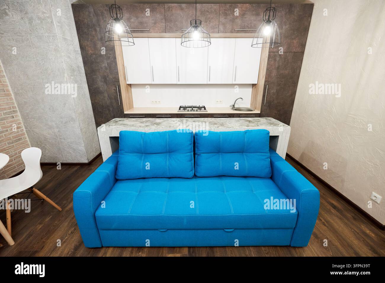 Blaues Sofa vor kompakter Küche mit Theke in moderner Studio-Wohnung. Stilvolles Open-Space-Interieur mit Pendelleuchten, Holzfußboden und urbanem Wohnkonzept Stockfoto