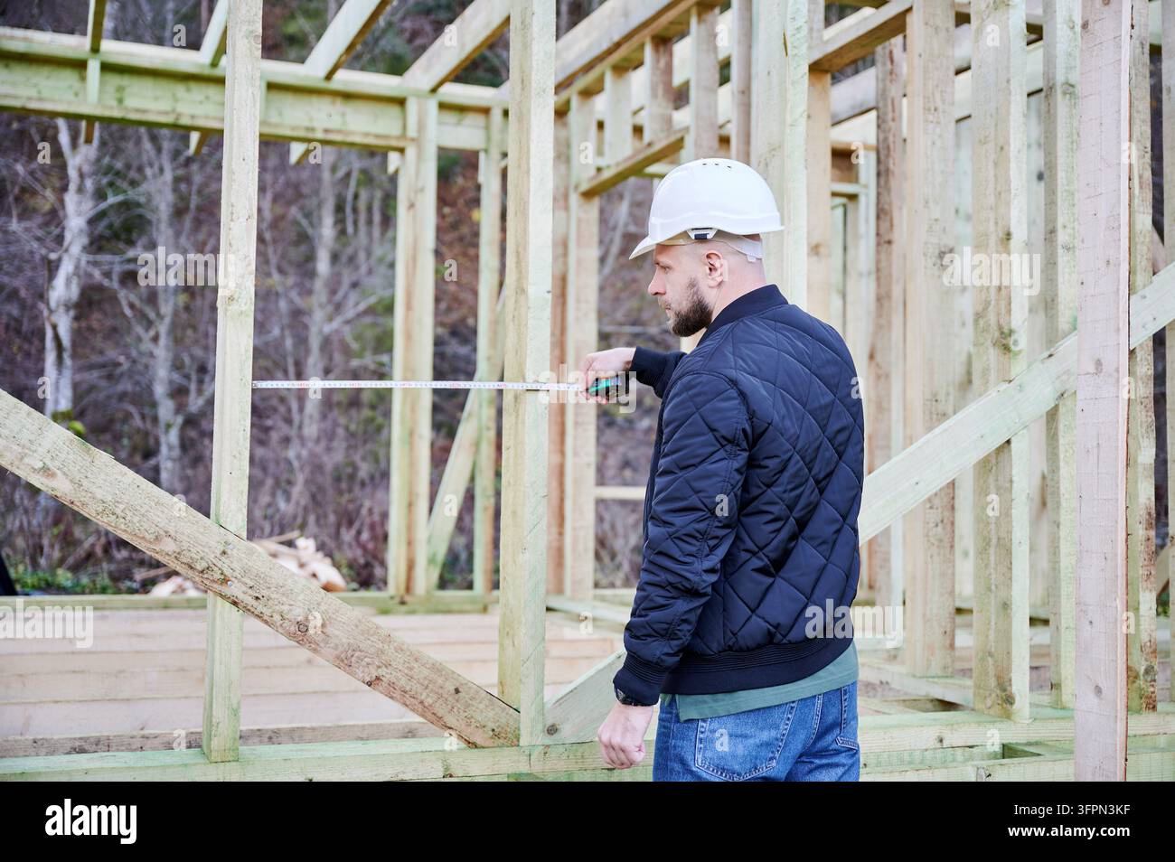 Bauarbeiter misst den Abstand zwischen Holzstützen mit einem Maßband im Rahmen des Hauses, überprüft den Abstand und die strukturelle Anordnung während des Bauprozesses Stockfoto