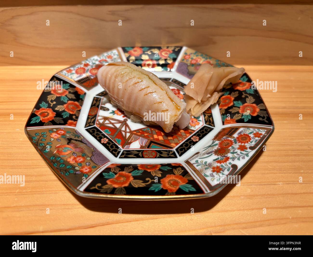 Ein gebratener Barfin-Flunder-Nigiri-Sushi, serviert auf einem traditionellen bunten Arita-Keramikteller auf einer Holztheke in einem japanischen Restaurant. - Smartphone-aufgenommenes Stockfoto