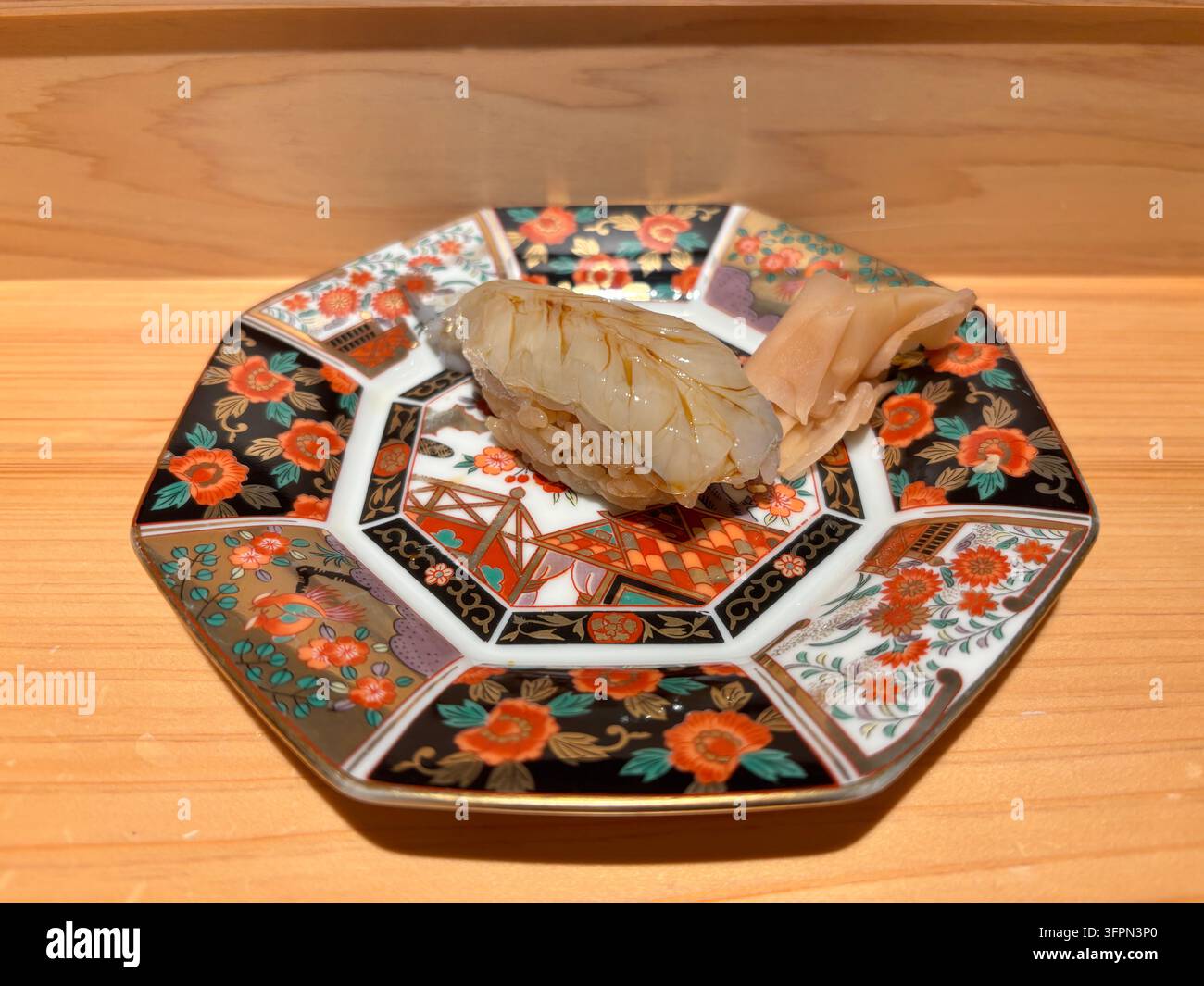 Ein glasierter Garnelen-Nigiri-Sushi, serviert auf einem traditionellen bunten Arita-Keramikteller auf einer Holztheke in einem Restaurant. - Smartphone-aufgenommenes Stockfoto