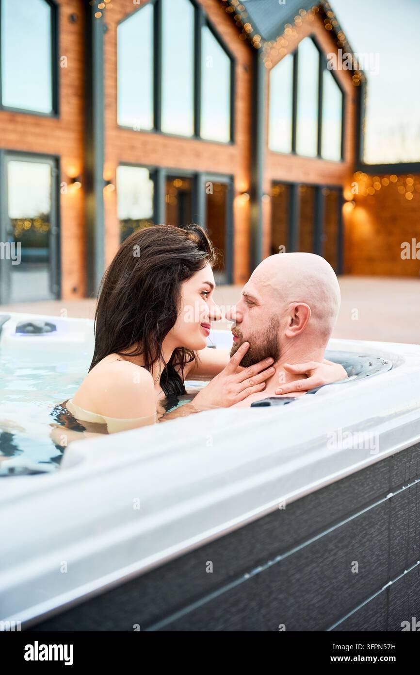 Paar genießt einen Whirlpool in der Nähe eines modernen Holzhauses mit festlichen Lichtern. Partner genießen einen gemütlichen Abend, Entspannung, Romantik und Urlaubsatmosphäre als Konzept Stockfoto