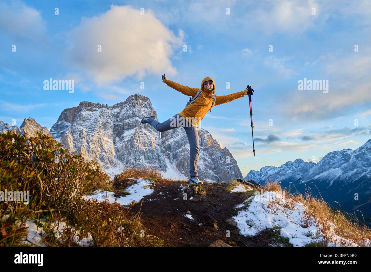 Fröhliche Frau posiert wie fliegend, während sie auf einem Alpenkamm balanciert, mit Trekkingstöcken neben schneebedeckten Dolomiten-Gipfeln in Italien, und veranschaulicht unbeschwerten Reisen, Outdoor-Wellness und abenteuerlichen Geist. Stockfoto