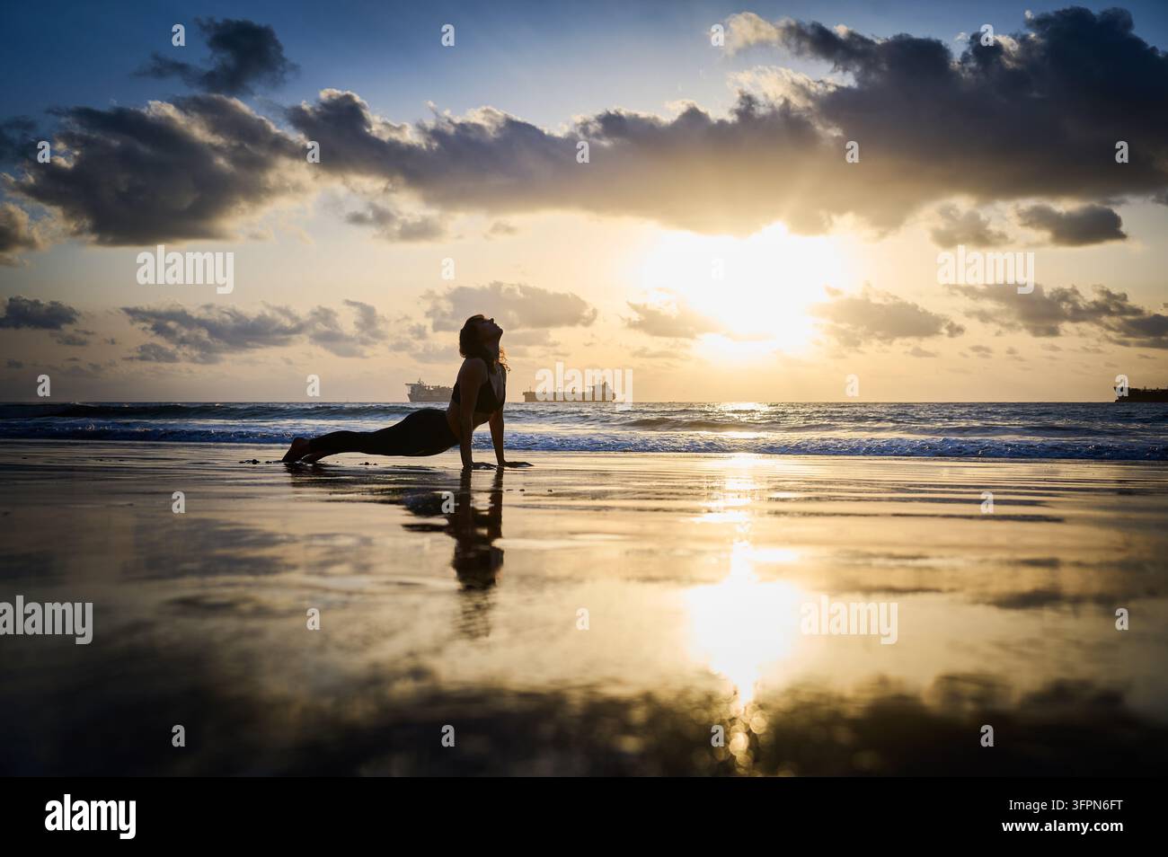 Frau führt im Sonnenaufgang auf Playa de Las Gaviotas, Teneriffa, die nach oben gerichtete Hunde-Yoga-Pose aus, streckt sich neben dem Atlantischen Ozean mit Frachtschiffen vor der Küste und warmen Reflexionen auf nassem Sand Stockfoto