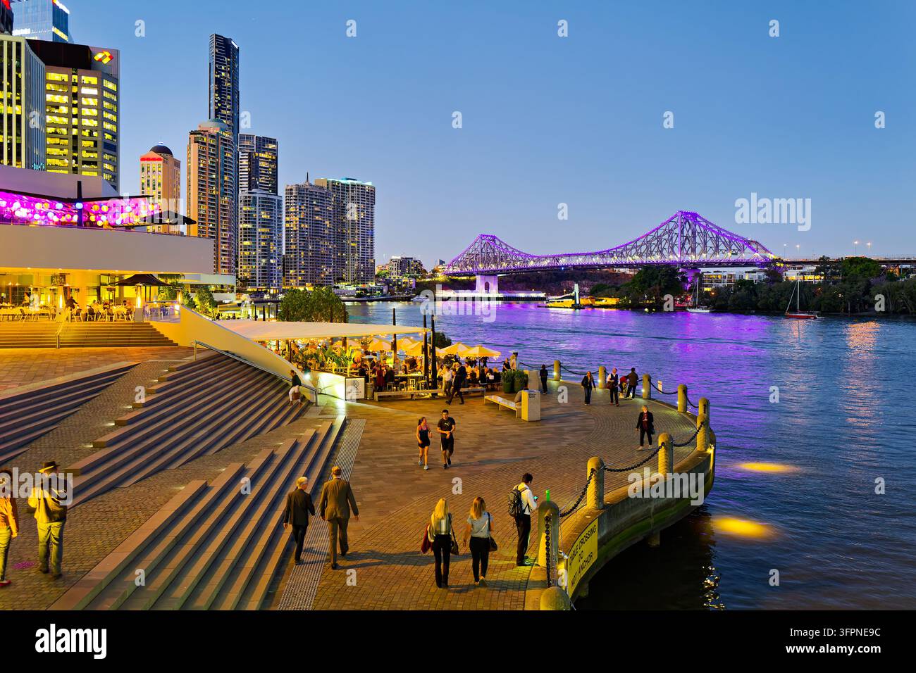 Fußgänger gehen an Außengastronomie am CBD-Wasserfront vorbei, während die Story Bridge in der Blauen Stunde lila leuchtet, Brisbane, Queensland, Australien Stockfoto