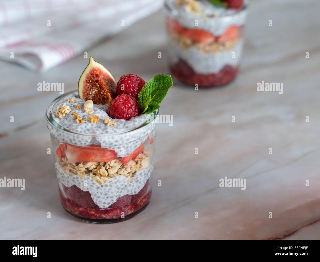 Individuelles geschichtetes Parfait mit Feigen, Himbeeren, Chiasamen und Granola, kombiniert mit einer cremigen weißen Schicht und frischer Minze für ein farbenfrohes Obstdessert. Stockfoto