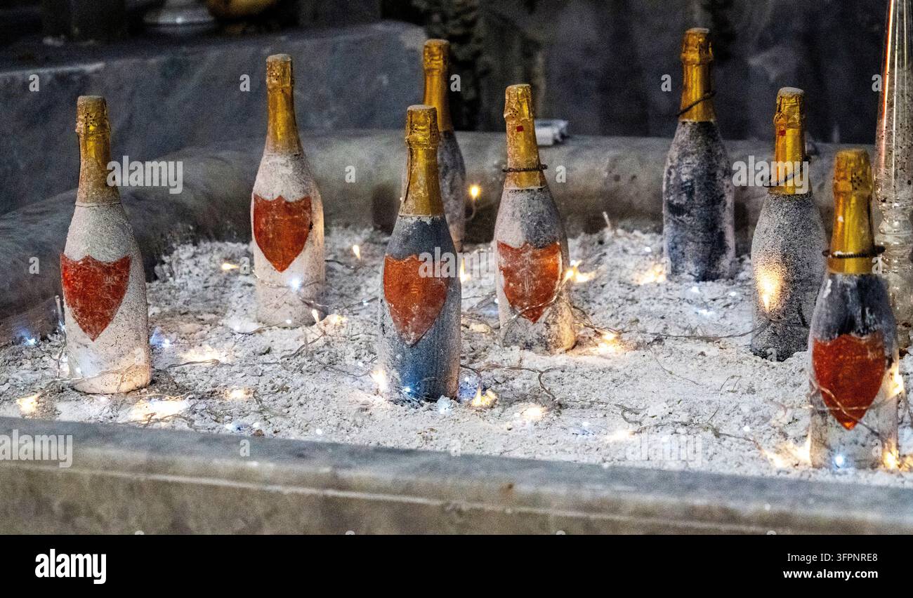 Kleine Champagnerflaschen mit goldfarbenem Folienverschluss und roten Herzetiketten stehen in Asche und glühenden Kerzenflammen an einem Schrein. Stockfoto