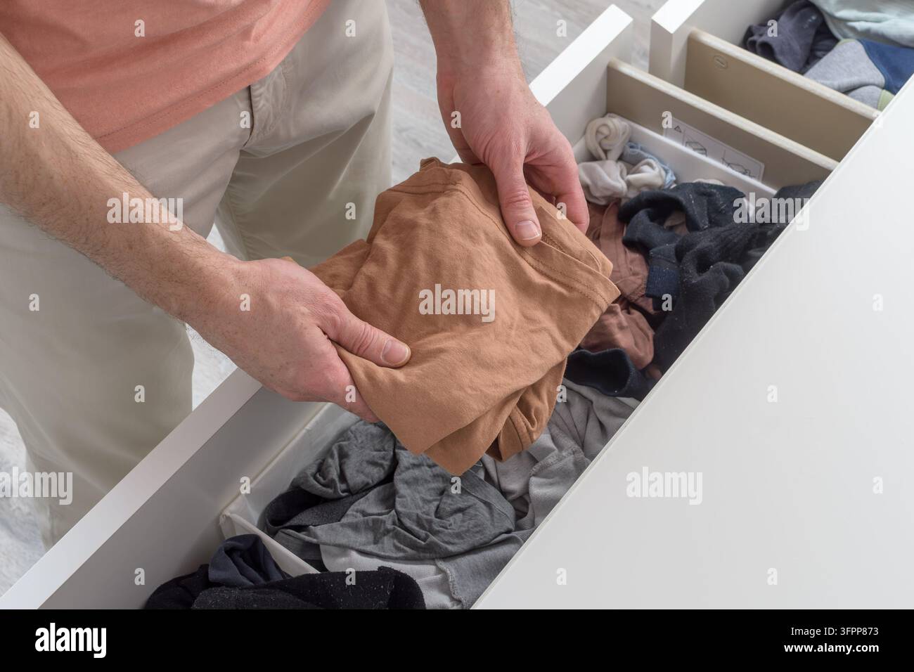 Mann zieht beiges T-Shirt aus einer überfüllten Kommodenschublade, die Aufräumung der домашних Kleidungsstücke zeigt alltägliche Routine und häusliche Verantwortung. Stockfoto