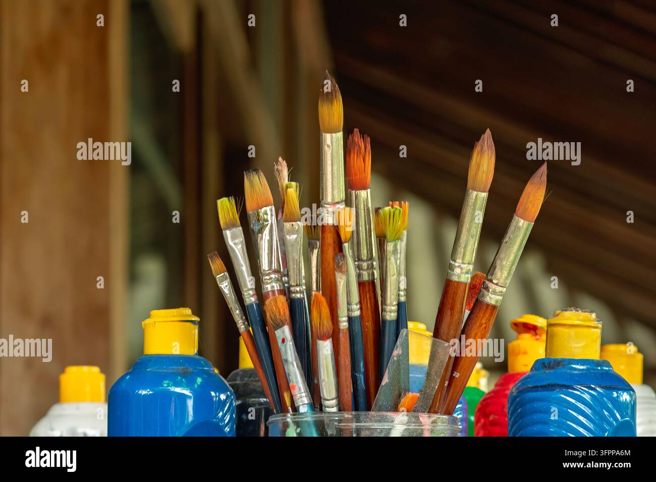 Ein Glas gefüllt mit Pinsel, umgeben von bunten Farbflaschen. Ein Künstlerarbeitsplatz unter einem Holzdach, bereit für Kreativität. Stockfoto