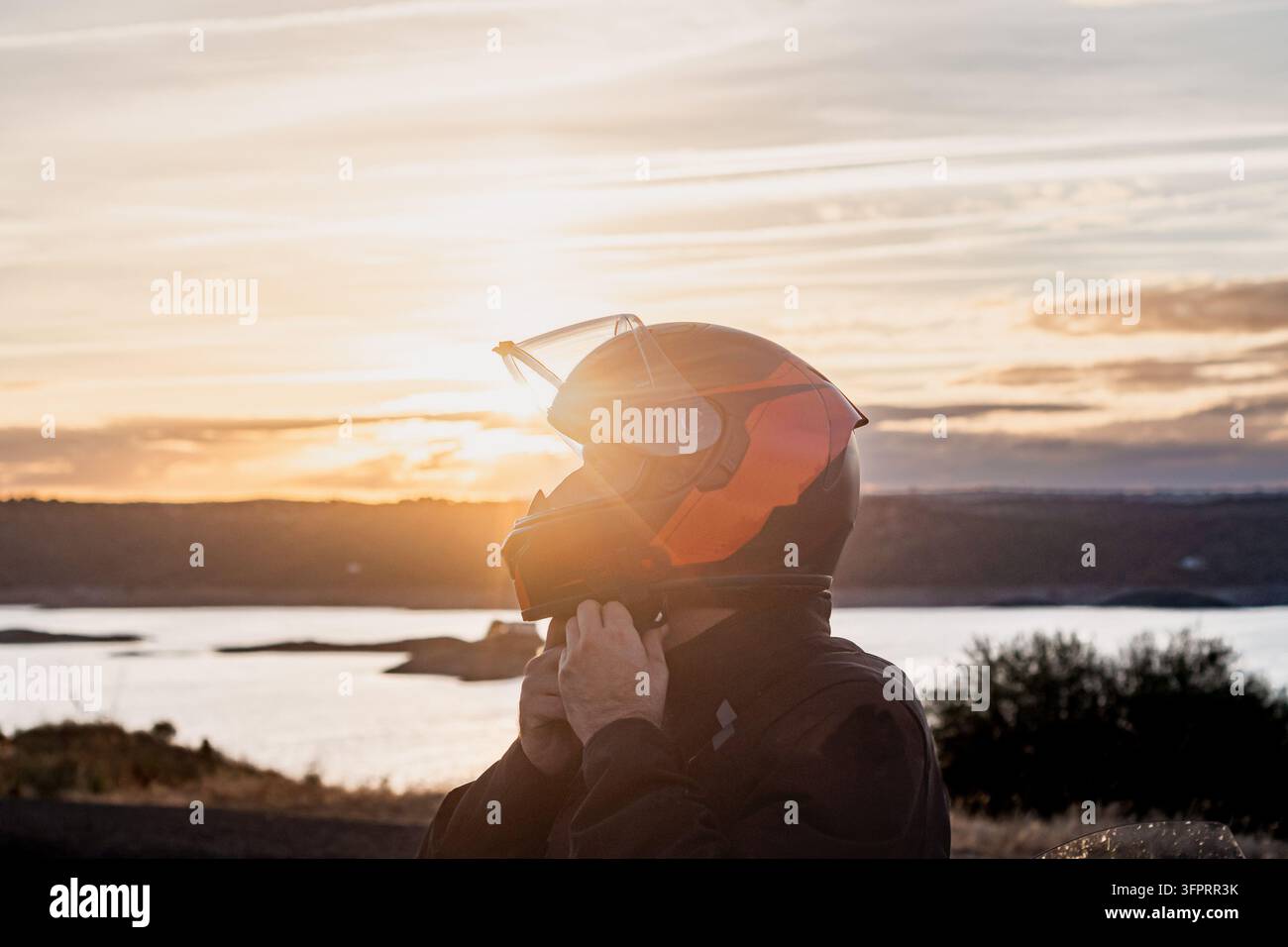 Profilporträt eines Motorradfahrers, der seinen Vollvisierhelm mit orangefarbenen Akzenten vor einem leuchtenden Sonnenuntergang und einer Stauseelandschaft in Extremadur befestigt Stockfoto