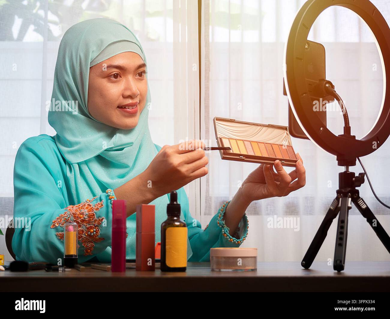Glückliche junge asiatische muslimische Frau, die von zu Hause aus einen Beauty-Vlog streamt, Online-Content-Erstellerin, die ein Parfümduftmittel aufträgt Stockfoto
