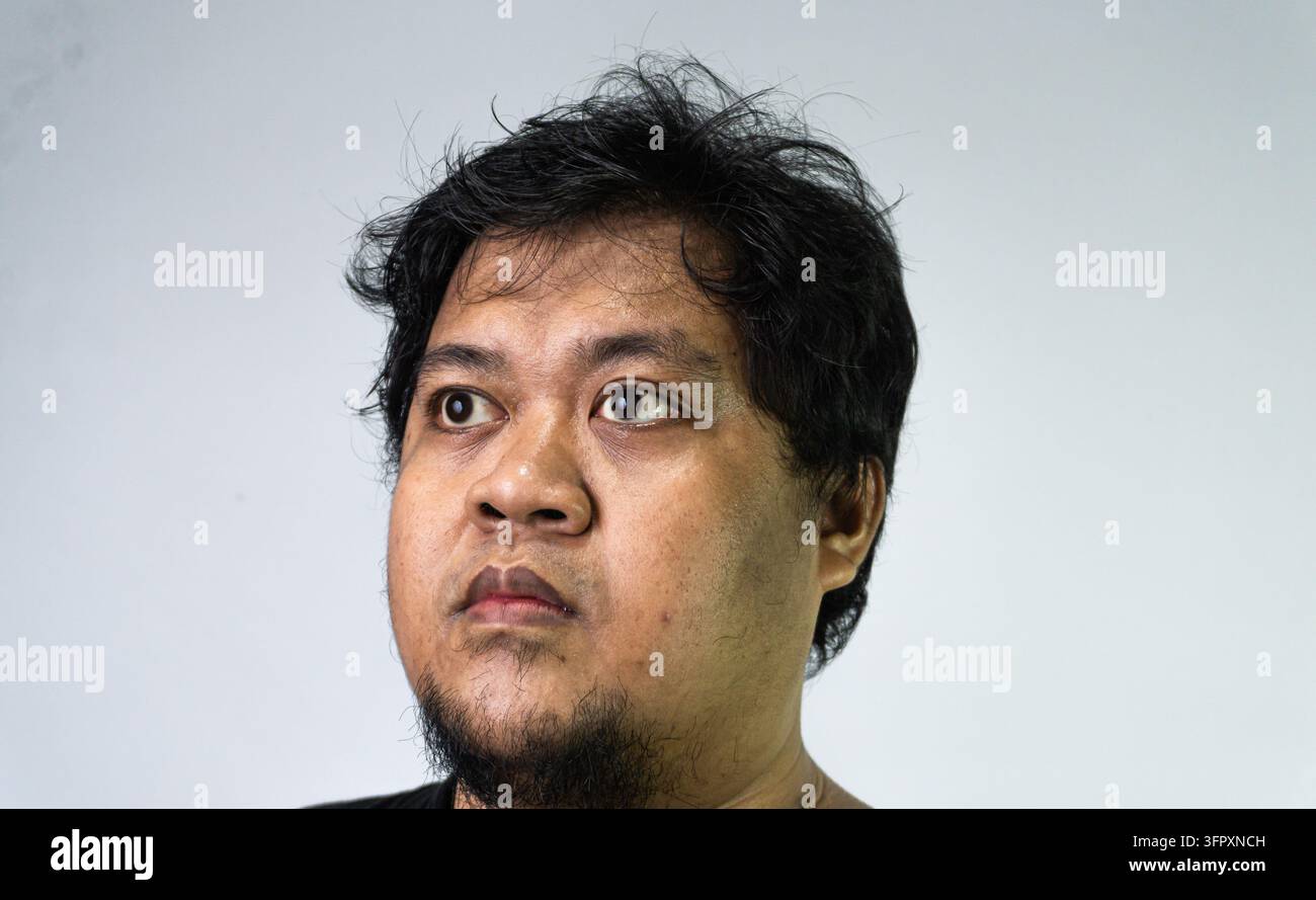 Ein junger asiatischer Mann schaut mit weit aufgerissenen Augen zur Seite vor einem hellen, minimalen Hintergrund und zeigt einen Ausdruck von Überraschung, Neugier oder Subtilität Stockfoto