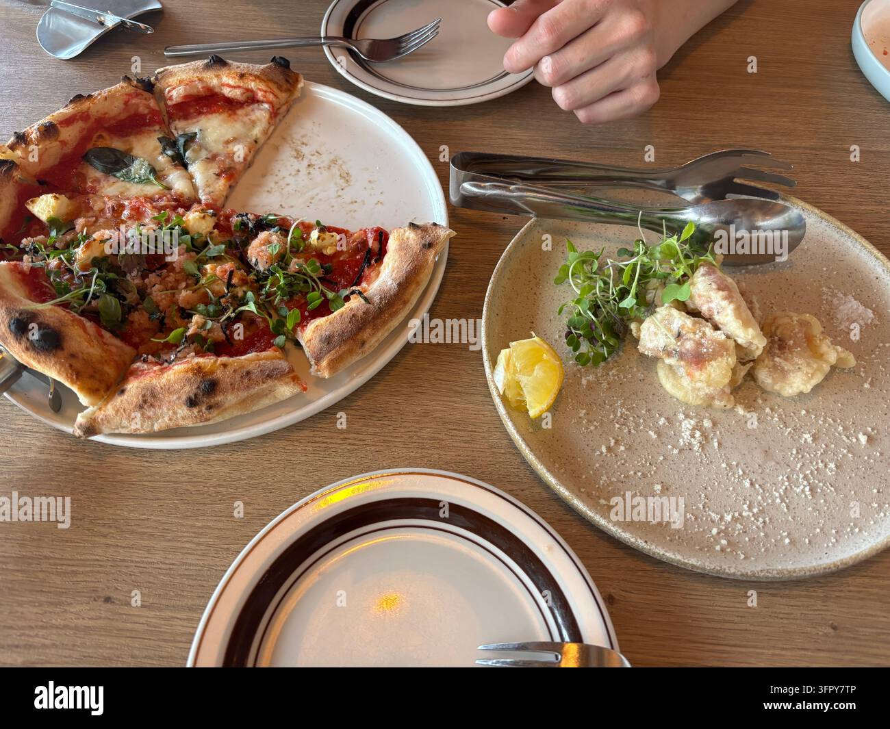 Frisch gebackene Pizzastücke, ein Beilagengericht aus frittiertem Fisch mit einer Zitronenscheibe, Teller zum Teilen und Servierzangen auf einem Holztischarrangement während einer Mahlzeit. - Smartphone-aufgenommenes Stockfoto