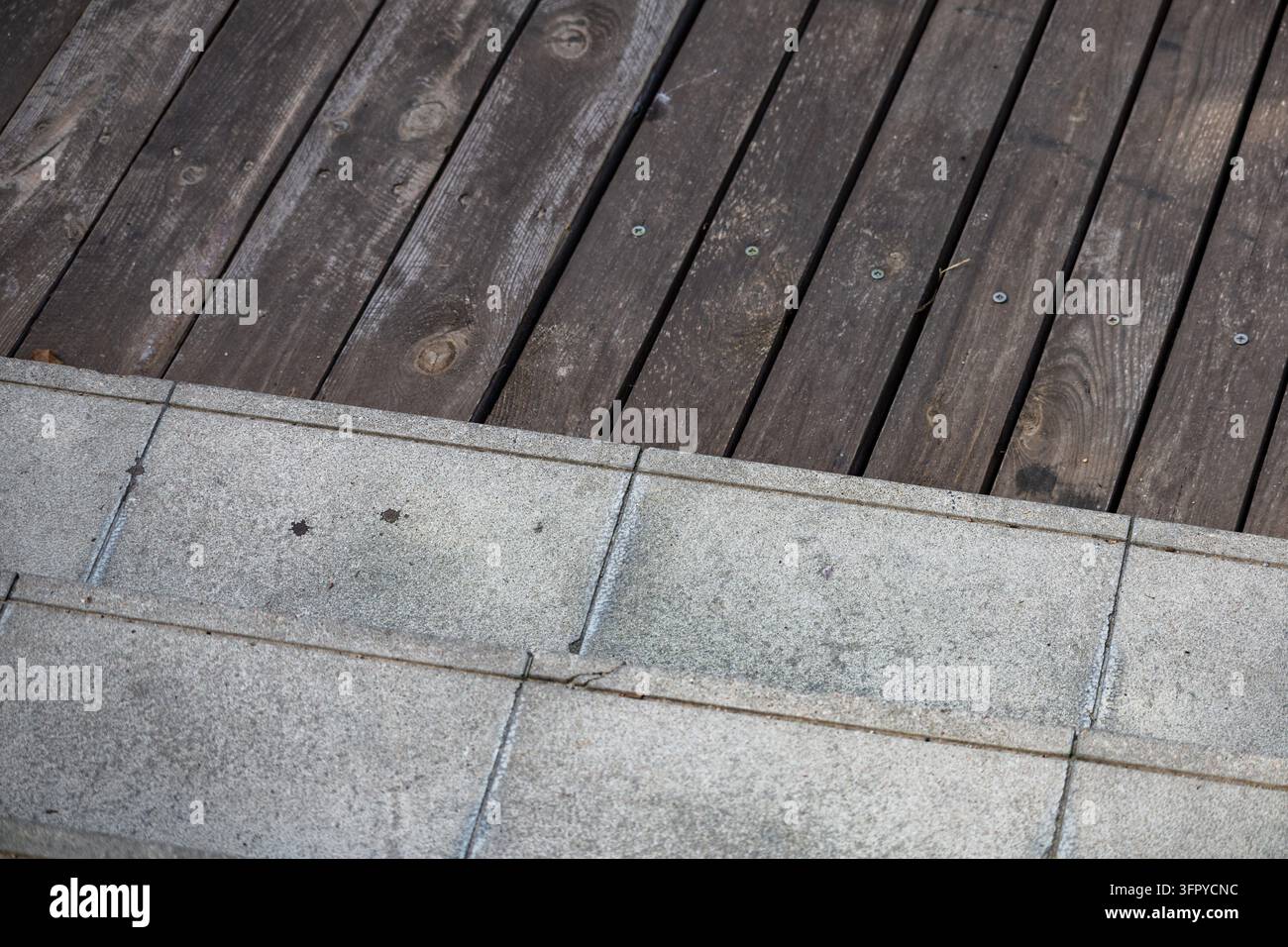 Graue Betonstufen treffen auf dunkelbraune, verwitterte Holzterrassenplanken und schaffen einen klaren, texturierten Oberflächenkontrast auf einer architektonischen Außenterrasse. Visi Stockfoto