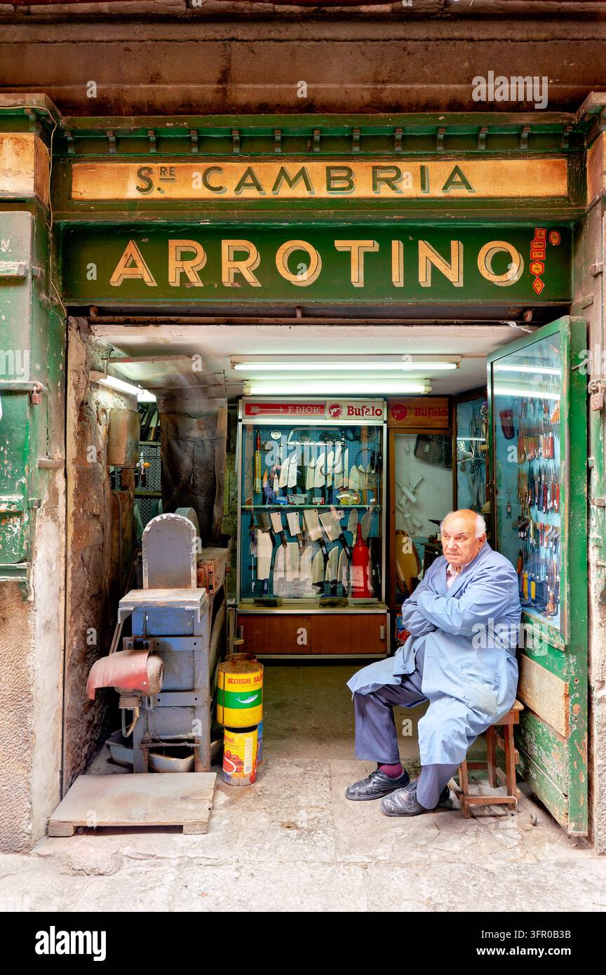 Ein alter Arrotino in einer blauen Arbeitsjacke sitzt auf einem Hocker an der offenen Tür einer traditionellen Messerschmiede in Siziliens Hauptstadt Palermo. Stockfoto