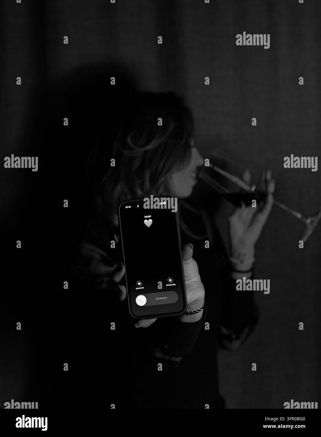Schwarzweißbild einer Frau, die ein Smartphone benutzt, während sie zu Hause ein Glas Wein hält. Stockfoto