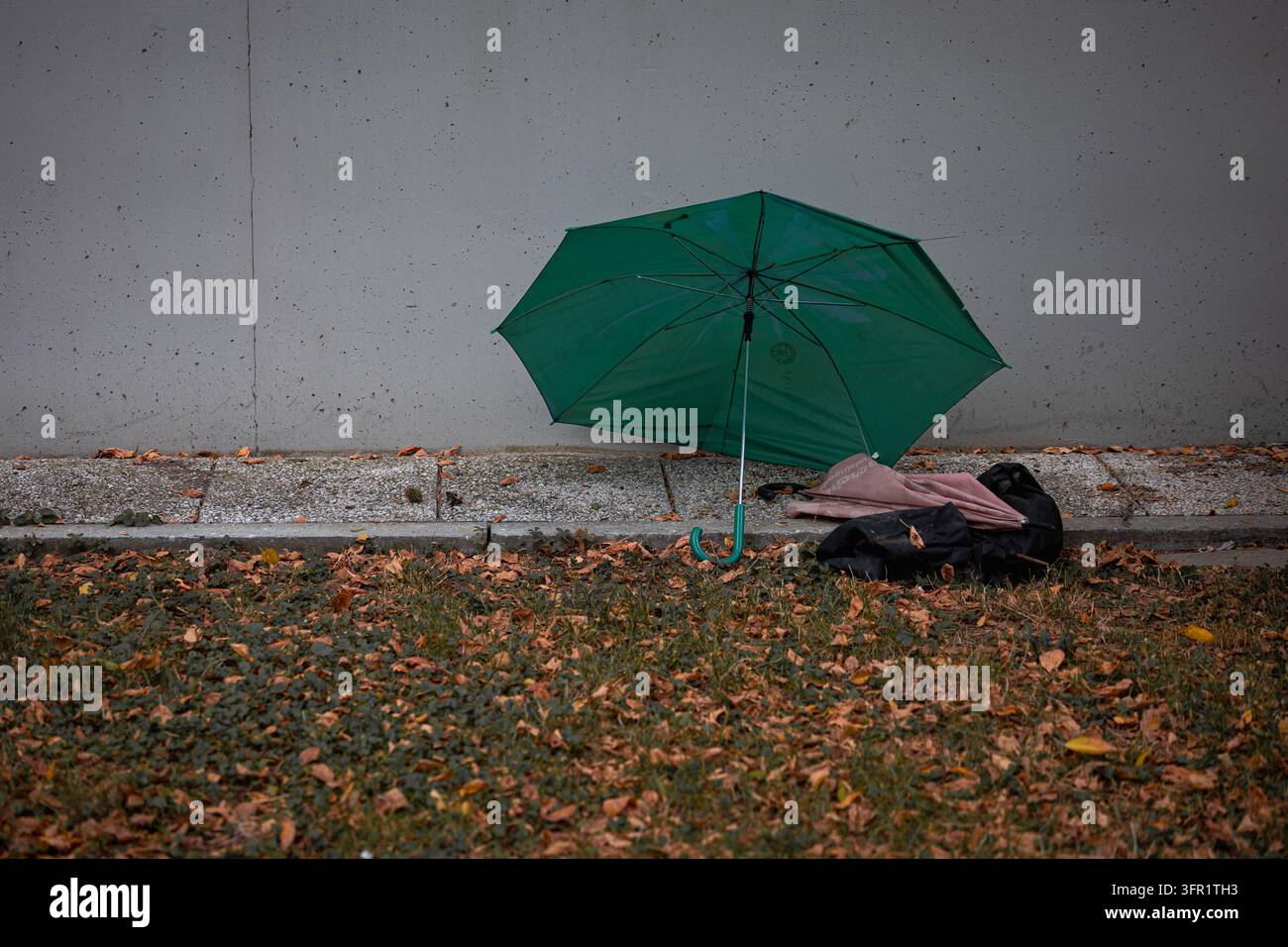 Verlassener grüner Regenschirm liegt auf dem Bürgersteig nahe einer Betonmauer an einem Herbsttag mit trockenen Blättern Stockfoto