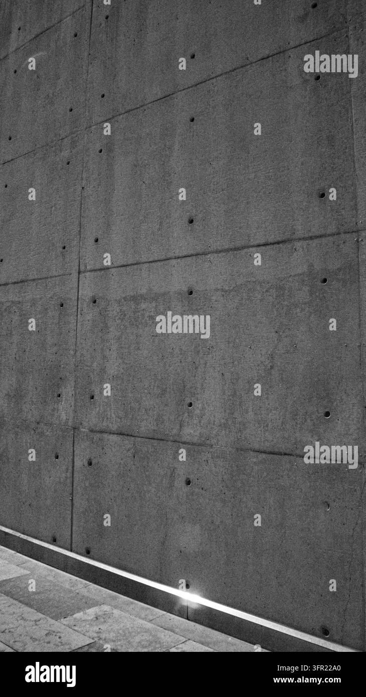 Eine Nahaufnahme, monochromatische Fotografie, die eine rohe Betonwand mit sichtbaren Nähten und kreisförmigen Vertiefungen zeigt. Eine lineare Leuchte verläuft entlang der Stockfoto