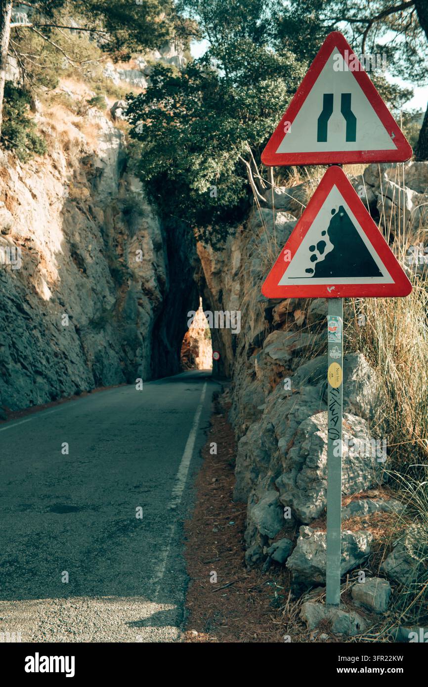 Eine niedrigwinklige, vertikale Aufnahme zeigt zwei rote und weiße Warnschilder – Straßenverengung und Steinschlag – die neben einer schmalen Straße in Mallorca, Spanien, stehen. T Stockfoto