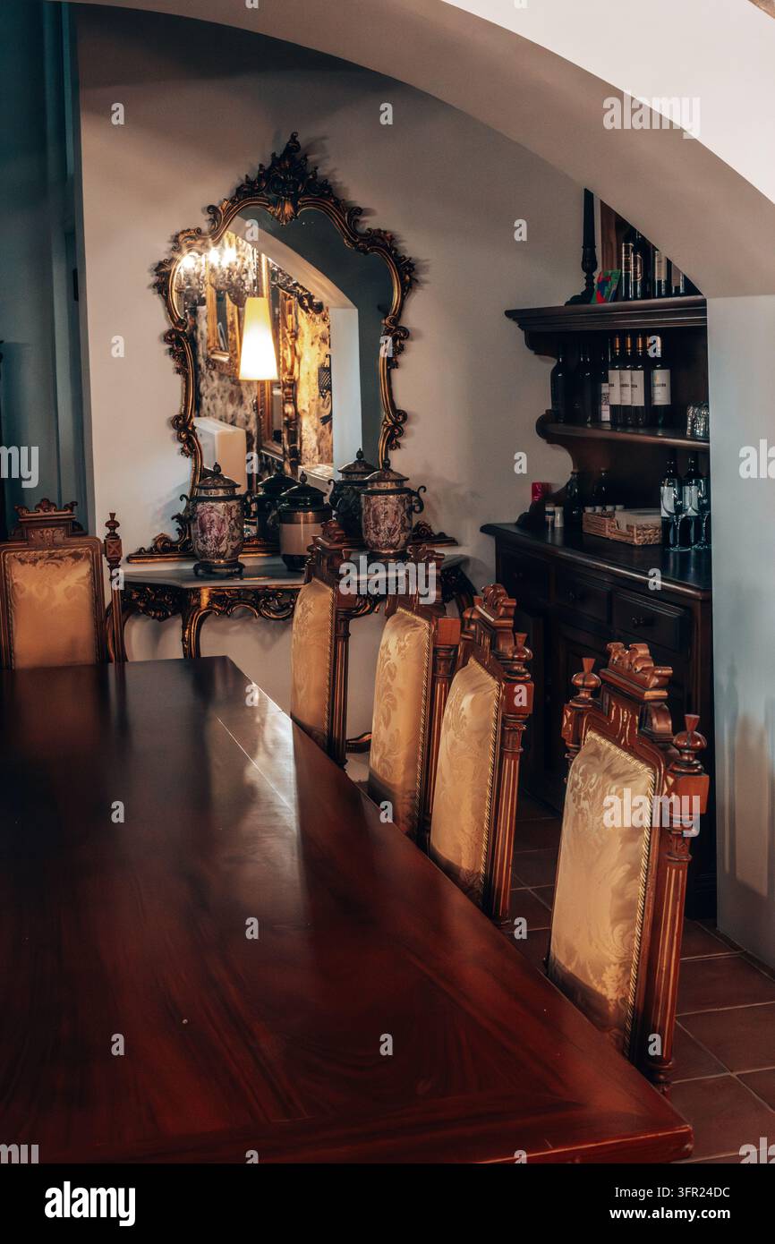 Ein elegantes Esszimmer-Interieur, das klassisches Design mit einem langen, dunklen Holztisch und kunstvoll gepolsterten Stühlen präsentiert. Ein großer, vergoldeter Spiegel reflektiert Stockfoto