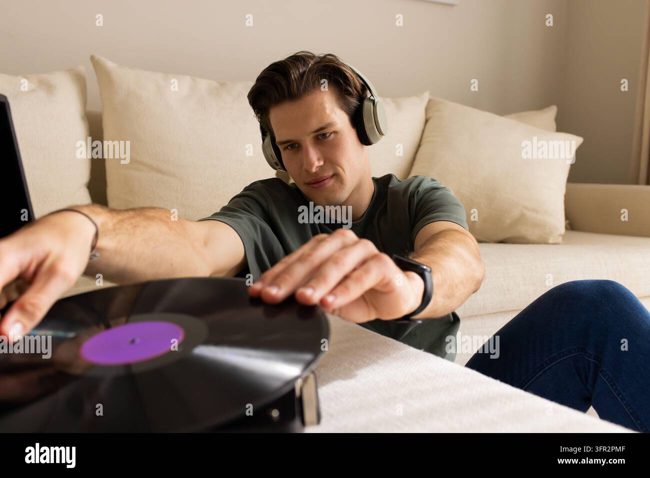 Mann mit Kopfhörern, grünes T-Shirt, der eine schwarze Vinylplatte auf einen Plattenspieler auf einem beigen Sofa legt Stockfoto
